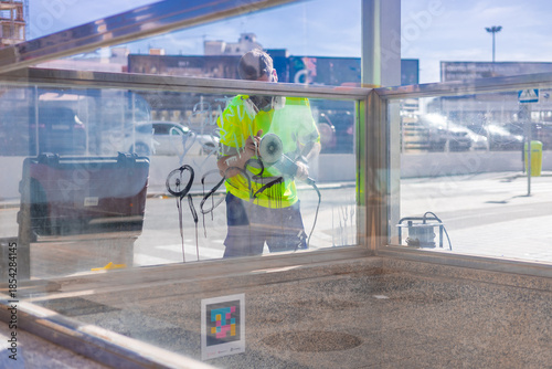A worker in a high-visibility yellow shirt uses a power tool to remove graffiti from a glass transit shelter in Valencia. They wear protective gear while sanding off black spray-painted markings.