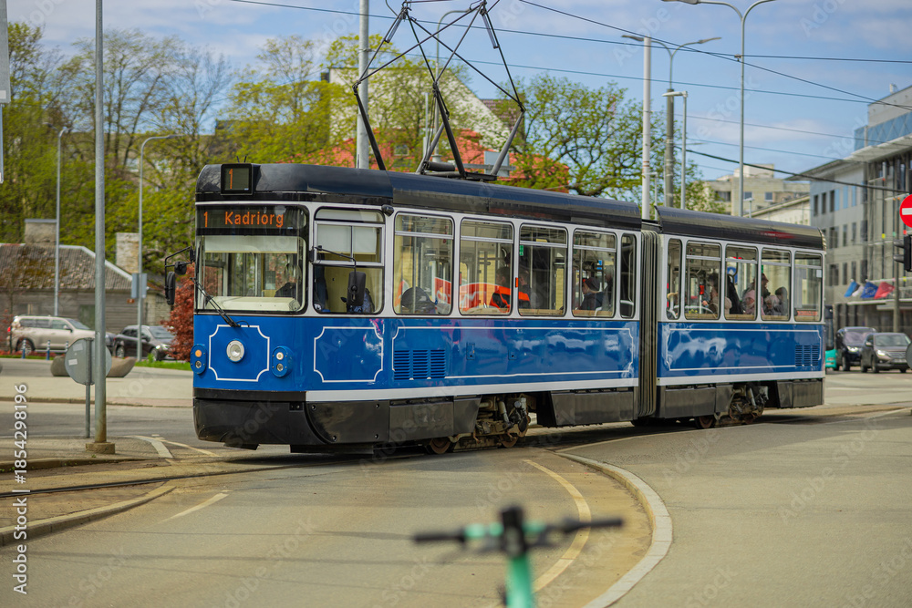 Naklejka premium A blue and white vintage-style tram travels along a curved track in Tallinn. The tram, headed for Kadriorg, moves through an urban intersection under a bright, overcast sky.