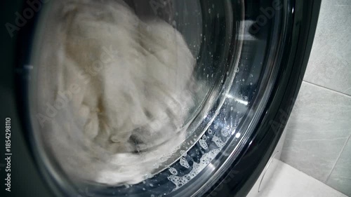 A close-up of old, yellowed gray laundry that was once white being washed in a washing machine. Bubbles of detergent drip down the glass window.