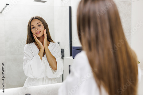 Pretty woman making face-lifting massage in bathroom, looking at mirror and smiling, using moisturizing face cream