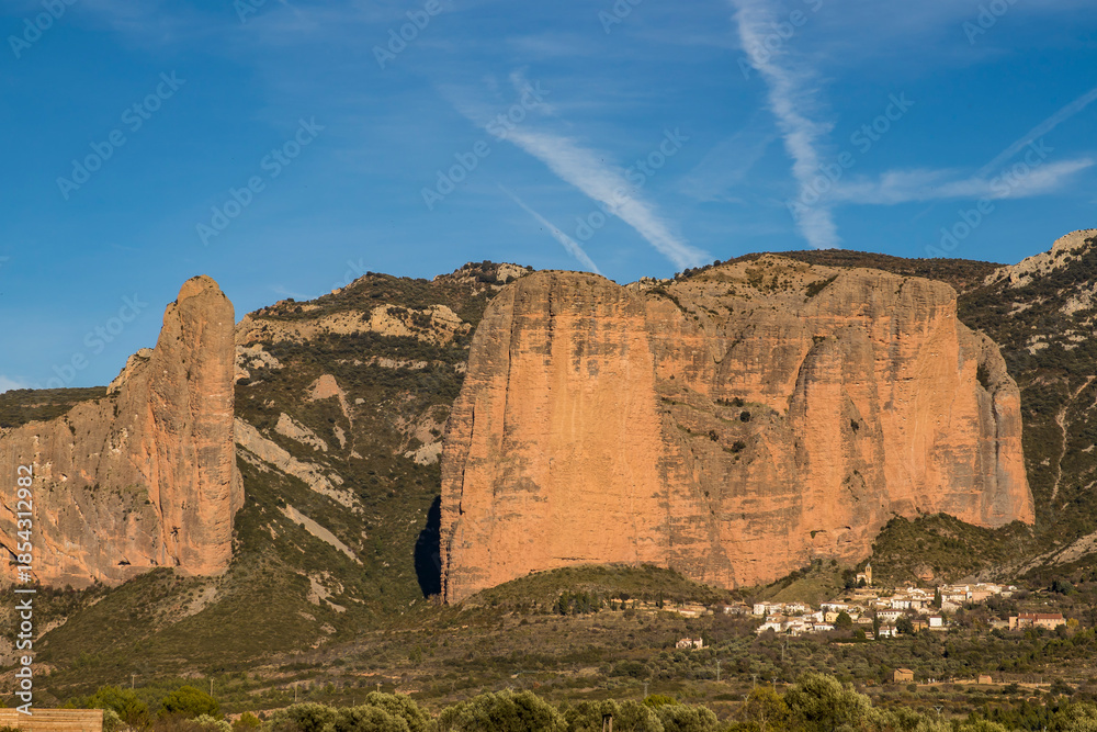 Fototapeta premium Olive Grove with the Mallos de Riglos Rock Formations and the Village of Riglos on a Sunny Day, Spain