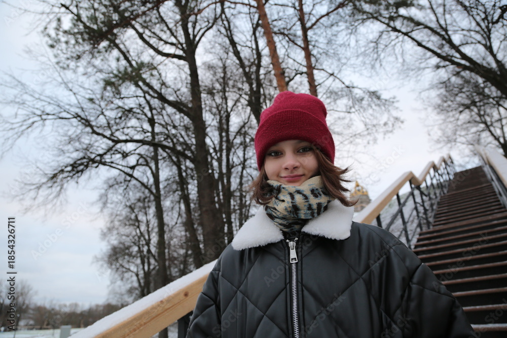 Fototapeta premium Happy fun smiling young woman walking in the city street winter park with trees in red hat and fashion black coat with copy space background