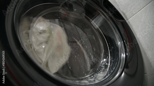 A close-up of old, yellowed gray laundry that was once white being washed in a washing machine. Bubbles of detergent drip down the glass window.
