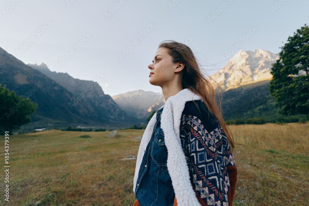 Naklejka premium Woman profile in mountains valley nature portrait outdoors landscape wearing patterned jacket with shearling, contemplative young traveler gazing across alpine meadow in serene sunlight.
