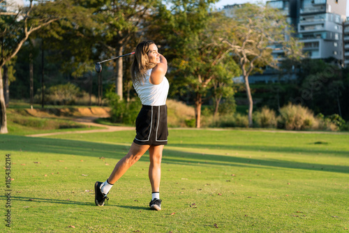 A latin athletic woman performs a powerful golf swing on a lush green course under golden hour sunlight