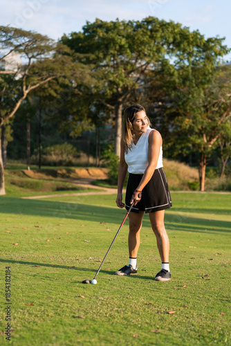 An hispanic woman stands on a sunny golf course holding a club near a golf ball, looking to the side before her swing
