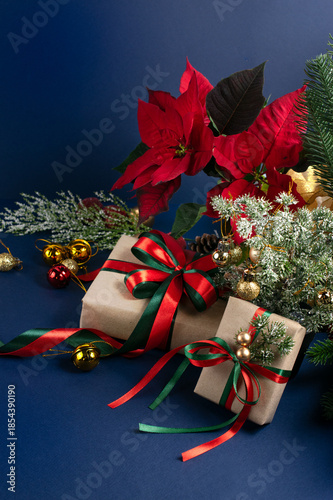 Christmas decor with handmade gift boxes, red poinsettia, decorative fir branch, bells and balls for tree decoraton on blue vertical background. New Year winter holiday composition with presents.