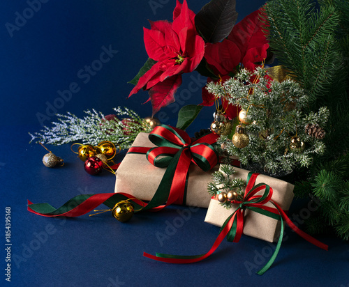 Christmas decor with handmade gift boxes with bow, red poinsettia, decorative fir branch, bells and balls for tree decoraton on blue background. New Year winter holiday composition with presents.
