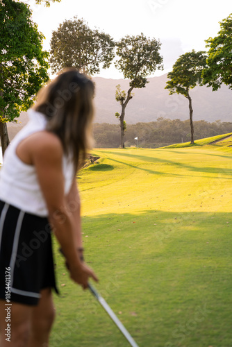 Latin woman preparing for a golf swing on a green course, enjoying outdoor sport and leisure activity during sunset