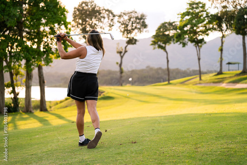 Back view of woman swinging golf club on a green course during sunset. Enjoying an active lifestyle outdoors, playing sport