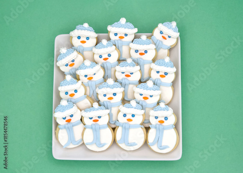Top above view of square porcelain plate filled with homemade sugar cookies snowmen with marshmallow fondant and royal icing. On a light green background.