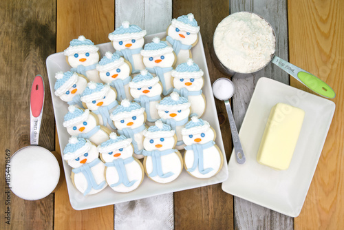 Top above view of square porcelain plate filled with homemade sugar cookies snowmen with marshmallow fondant and royal icing. Butter, flour, sugar and sale on wood table