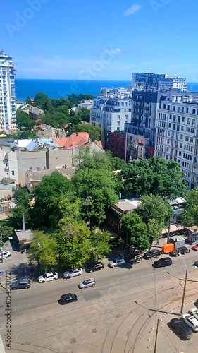 Cityscape. Tall buildings, moving cars, asphalt road, green trees, walking people, city street, sea and blue sky on a sunny summer day. City life. City landscape. City on sunny summer day. Vertical