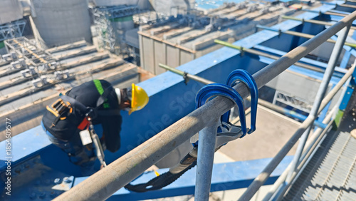 Safety lifeline hook attached to a steel railing at a high construction site, with a worker wearing helmet and harness blurred in background, emphasizing fall protection and workplace safety.