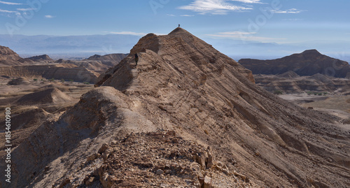 A sharp mountain ridge rises above wide and open valley of Nahal Zinin or Zanin in the Negev. A pair of tourists stands on a cliff with a panoramic view of the valley. Panoramic desert landscape.