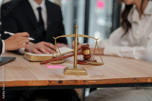 A brass scale of justice and a wooden gavel placed on a desk while professional lawyers discuss legal cases and sign documents in the background.