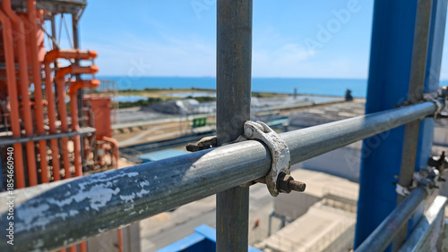 Close-up of metal scaffolding pipes and couplers secured with bolts at an industrial construction site, with offshore platform and blue sky in background, highlighting structure, safety, and engineeri