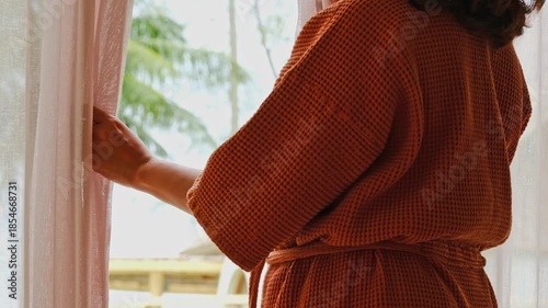 The woman was at the window in the bedroom. She opened the curtains on the window. In the morning and she looks at the view of mountains and trees at sunrise.	