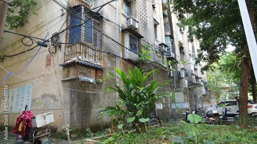 An older apartment complex in China with weathered concrete buildings, wiring, metal window grilles, and overgrown weeds in communal area. An aging residential neighborhood lacking maintenance