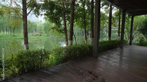 A quiet resting space within an urban park, featuring a wooden deck walkway shaded by trees and overlooking calm water and lush greenery. Contemporary public green space in Ningbo, China.