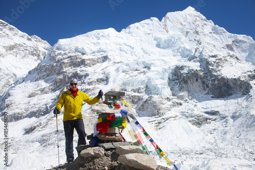 Adventurer in bright yellow jacket stands proudly beside colorful prayer flags at a stone monument, surrounded by majestic snow-capped peaks in a breathtaking mountain landscape.