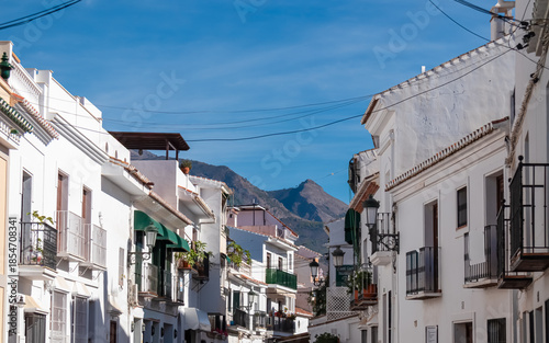 A narrow street lined with whitewashed houses and green awnings leads toward rugged mountains under a blue sky in Frigiliana, capturing the charm of a traditional Andalusian village in sunny Spain.