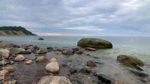 Rocky shore along calm sea with scattered stones and large mossy boulder under overcast sky near distant coastline. Quiet natural seascape showcasing rugged coast and tranquil waters.