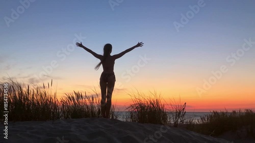 Silhouette of a person on a sandy dune at sunset. Arms outstretched signaling joy and freedom. Calm ocean horizon glows with warm colors creating peaceful inspiring mood.