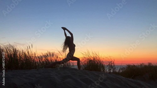 Silhouette of person practicing yoga on sandy beach at sunset with tall grasses and calm horizon. Tranquil fitness scene focused on balance flexibility and relaxation.