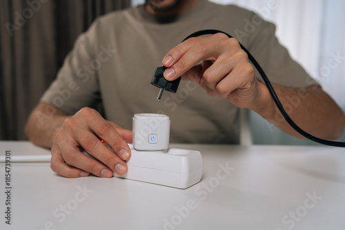 Photos Man holding power plug, connecting it to smart socket inserted in power strip, demonstrating modern smarthome technology for energy management and automation, close-up