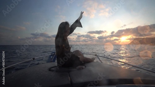 Woman boat sunset, young female traveler raising arms on a speeding motorboat enjoying the beautiful orange ocean dawn