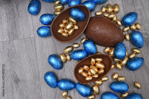 Pile or group of different sizes of colourful foil wrapped chocolate easter eggs in blue, and gold. Large halves of a brown milk chocolate egg have mini eggs inside, on a grey wooden background.