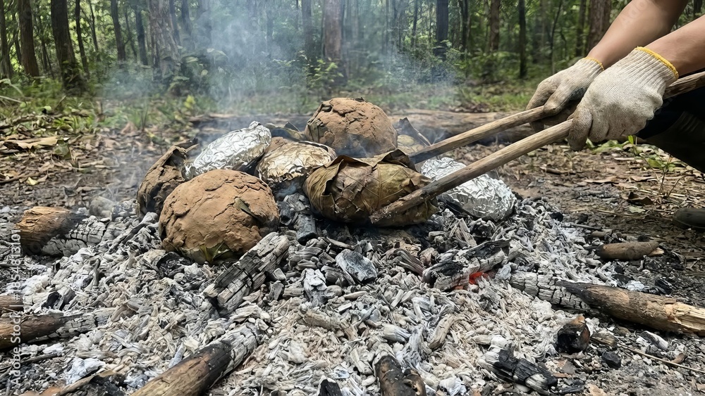 Obraz premium Person wearing gloves using tongs to carefully turn food packets wrapped in foil and leaves, baking in hot ashes and embers from a campfire in a forest