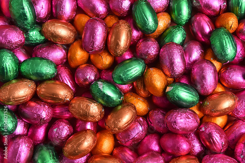 Large & small pink, green, orange and bronze foil wrapped chocolate easter eggs, against a peach orange background.