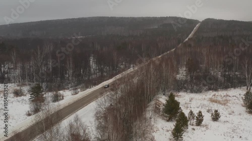 Aerial view of cars and a snowplow driving on a road cutting through a vast, snow-covered forest during a gloomy winter day