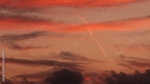 An airplane flies through a colorful sunset sky. Contrails are visible against the vibrant clouds, creating a striking contrast with the warm hues.