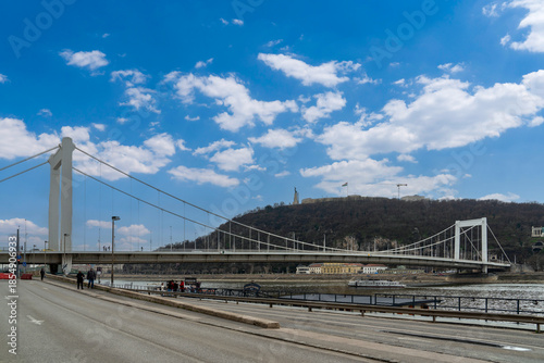 Photography Budapest, Hungary - March 15, 2025: A white suspension bridge, the Elisabeth Bridge, spans the Danube River in Budapest, with Gellért Hill and the Liberty Statue visible on the right under blue sky