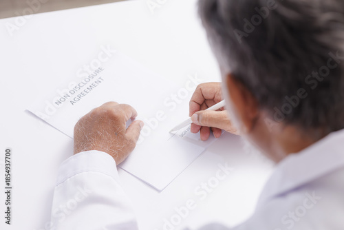 Person signing nondisclosure agreement at white desk, focusing on act of signing. setting is professional, with close up view of hands and document, emphasizing confidentiality