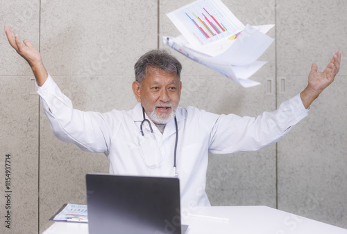 Doctor in white coat joyfully throws papers in air, sitting at desk with laptop, expressing relief or celebration. background is plain wall, suggesting professional setting