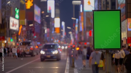 Vertical green screen billboard mockup on a busy downtown street at night. Blurry city lights, traffic, and pedestrians in the background.