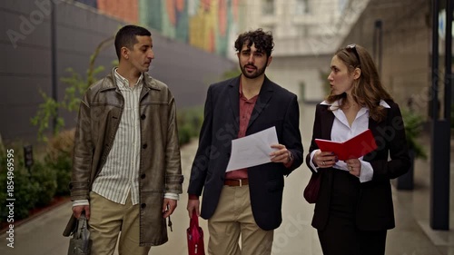 Business people, two men and one woman, collaborate and discuss a project while walking outdoors. Their dynamic interaction highlights modern teamwork and remote work flexibility in an urban setting.
