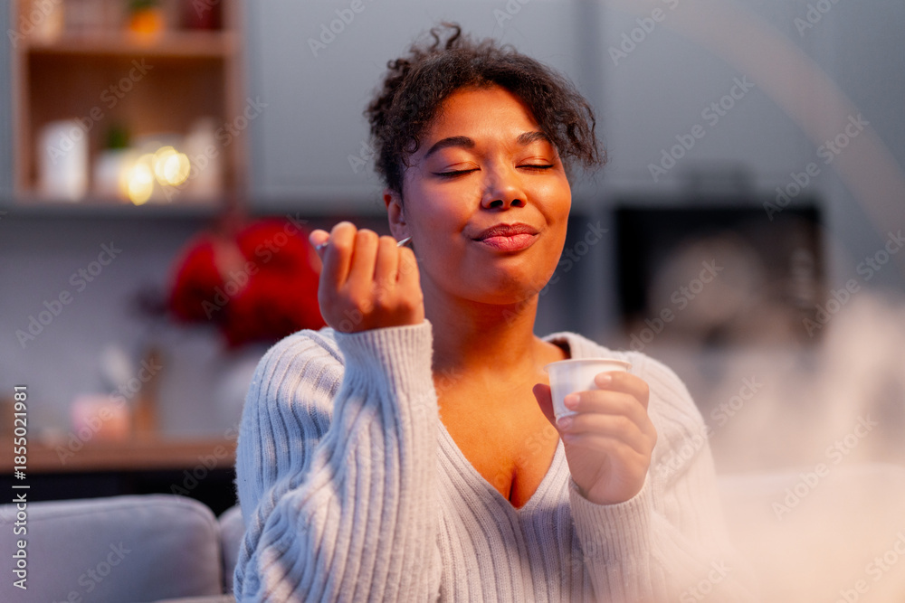 Fototapeta premium Young woman savoring a snack, finding peace and comfort in her indoor lifestyle