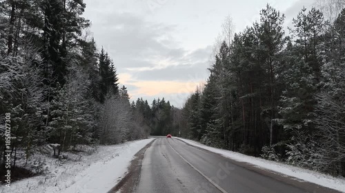 An asphalt road with markings, signs and sandy roadsides runs through a mixed forest. Snow is lying on the ground, roadsides and tree branches Cars are driving Clear winter evening and colorful sunset