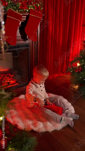 A little boy is sitting by the fireplace, opening a Christmas present. Christmas gift concept.