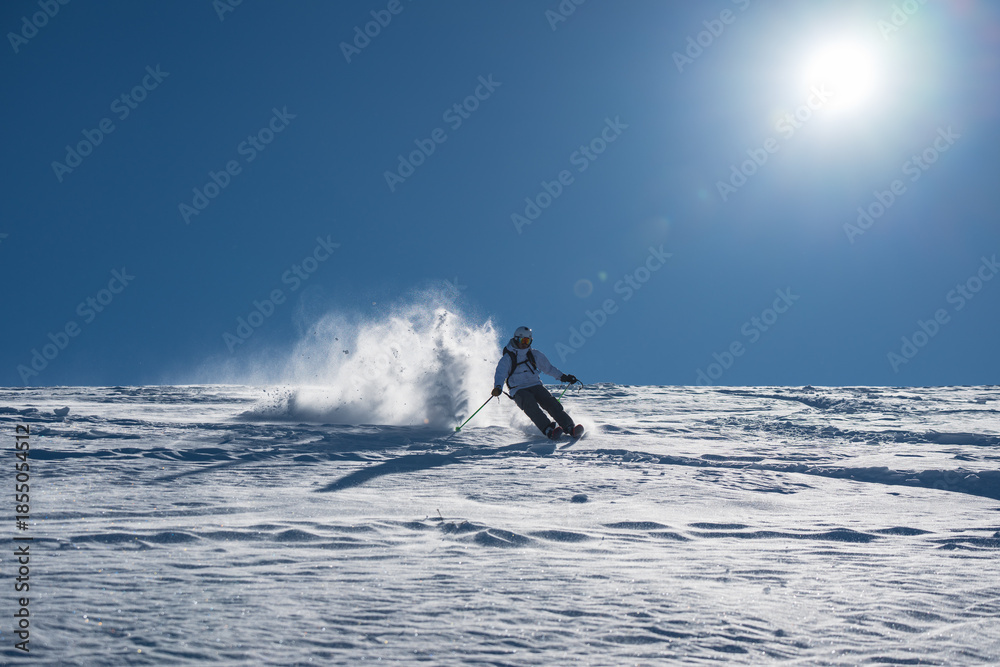 Naklejka premium Skier skiing off piste through fresh powder on a sunny mountain slope, kicking up snow as she descends, creating a dynamic and energetic winter sports action scene under a clear blue sky