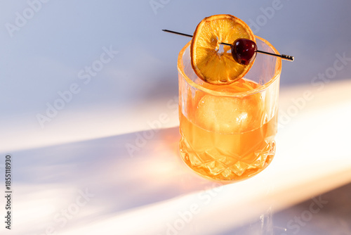 Close up of a manhattan cocktail served with one large ice cube on a reflective white surface with dual lighting tones and high contrast lights adding drama.
