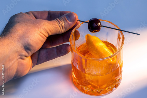 Close up of a hand placing the cherry on top of a manhattan cocktail served with one large ice cube.