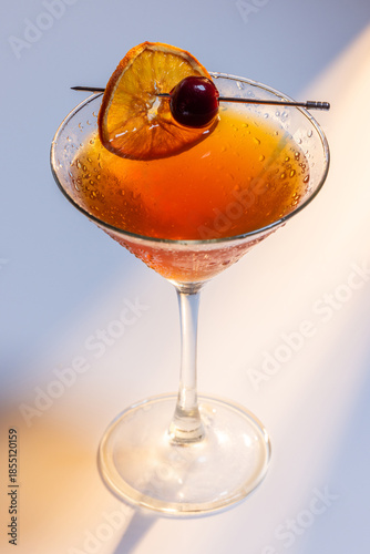 Beautiful amber cocktail, a Negroni or manhattan served up on a white surface with dramatic lighting.