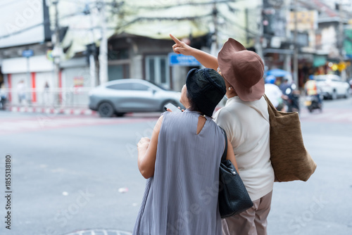 Senior asian women travelers standing on city corner pointing direction while checking smartphone representing friendship curiosity exploration and retirement travel lifestyle