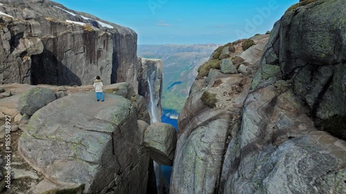 Woman standing on Kjeragbolten boulder above Lysefjord Norway. Drone shot shows a lone hiker balancing on the famous rock wedged between cliffs, overlooking the dramatic fjord landscape.